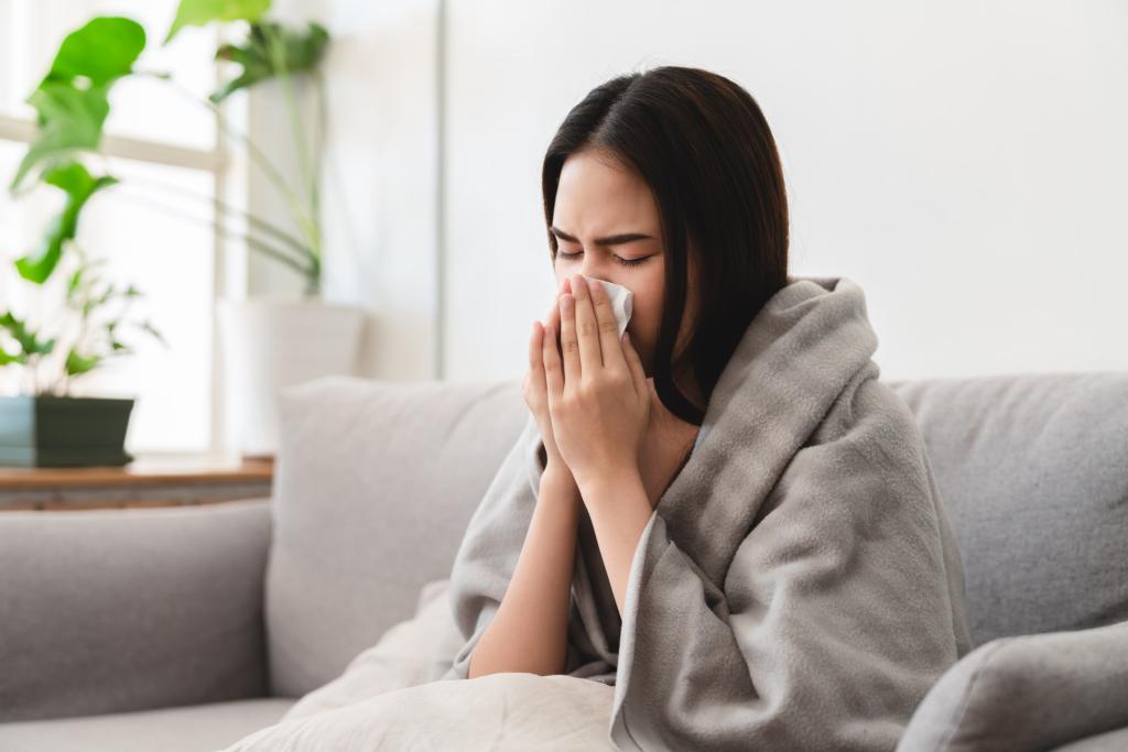 Sick young woman sneezing into tissue paper while covered with blanket sitting sofa living room.