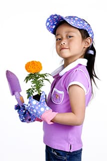 young girl wearing pink gardening shirt and jeans holding a pink trovel and orange flower,