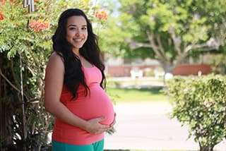 Pregnant women wearing a pink maternity shirt and turquoise pants holding/support her belly posing next to a wall of flowers