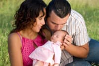 Man and woman couple holding a small baby girl in a pink dress, the family is sitting down in a field of grass.